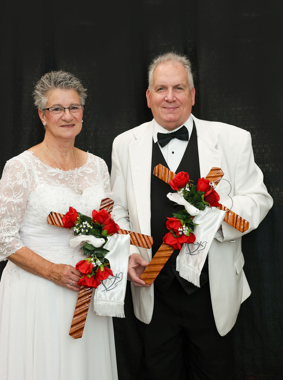 Man and woman dressed in formal, primarily white attire, holding crosses with roses and a dove.