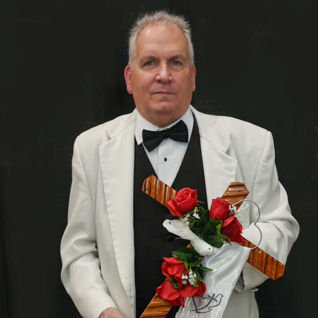 Man in suit and white jacket holding a wooden cross covered in roses.